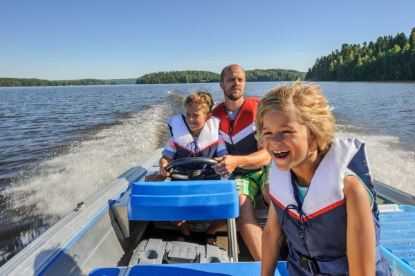 Father and sons out boating together. The father lets one of the boys steer the boat. They're wearing life vests. In the background there's forested shore line, water, and blue sky.